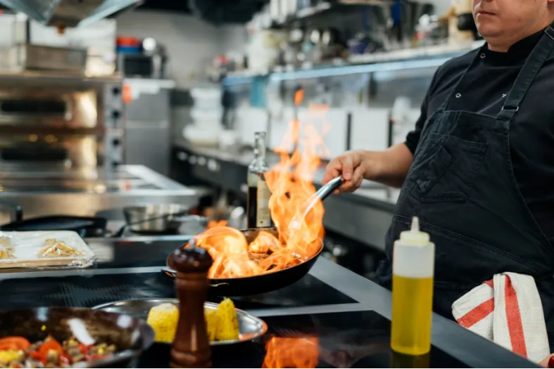 Flambé cooking in a professional kitchen with flames in a frying pan, chef preparing a dish with fire, restaurant culinary skills, professional kitchen setting.