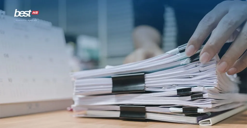 3bagiantantangan BEST Software Indonesia Powered by ProInt Unorganized stack of paperwork and documents on wooden desk with a person handling files representing business administration software document management or workflow automation by BEST Software Indonesia